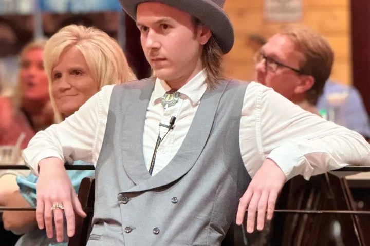 Person in grey suit and top hat leaning on a railing indoors.