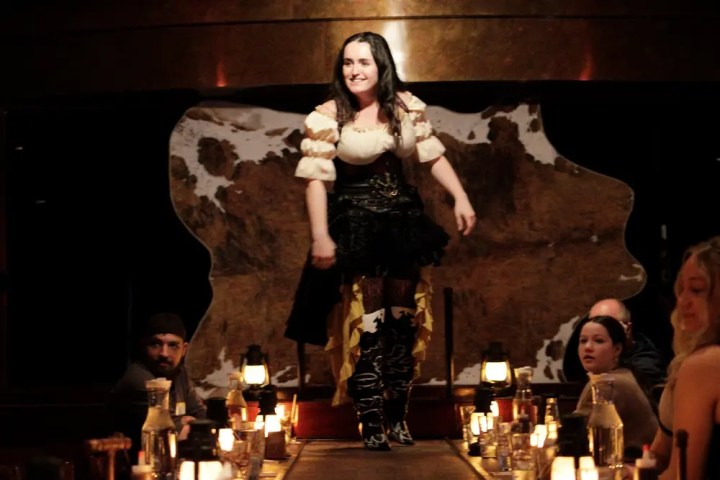 Woman in costume walking on table with people seated around.